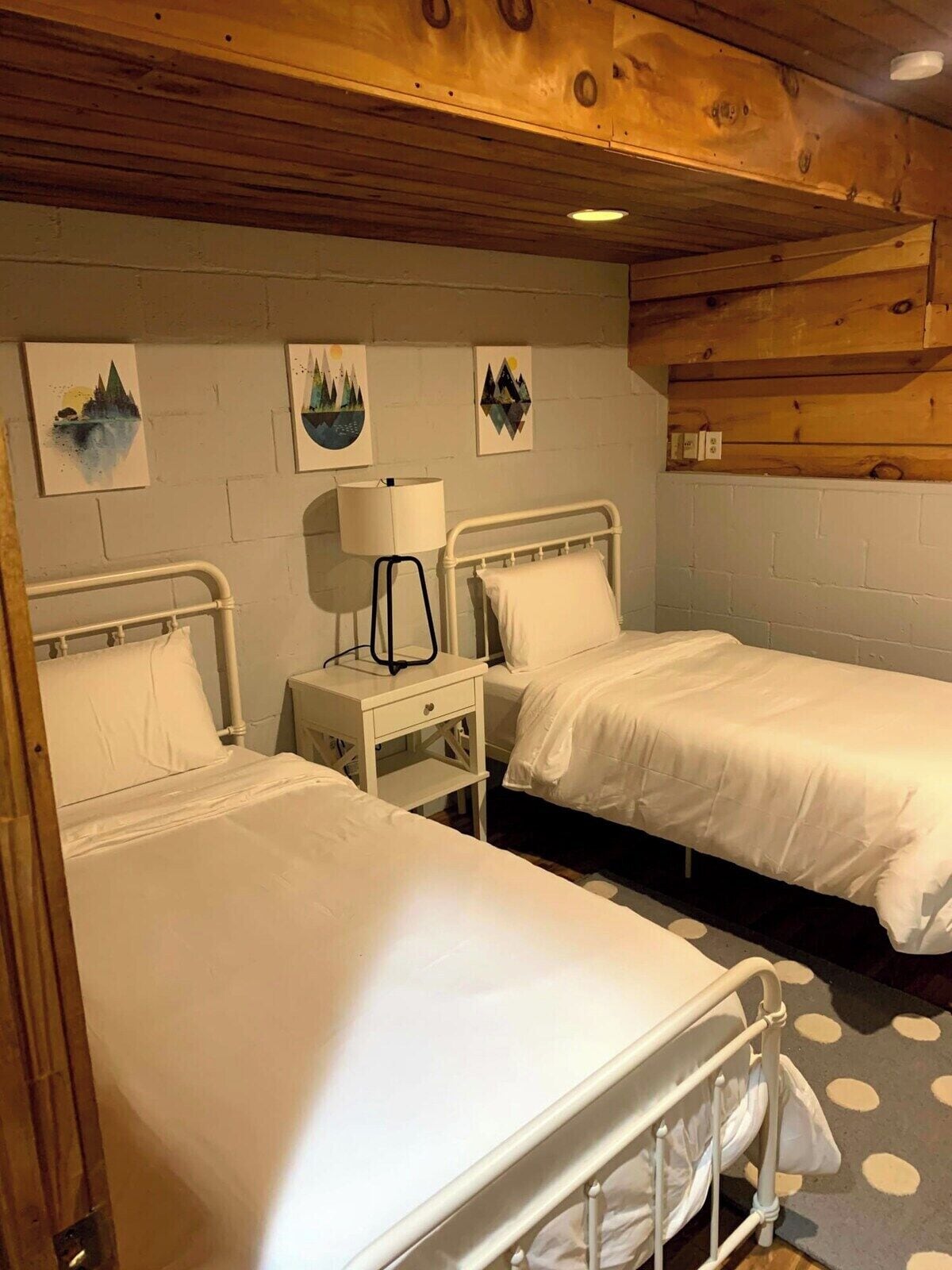 Una habitación con dos camas, una mesita de noche con lámpara y cuadros en la pared.