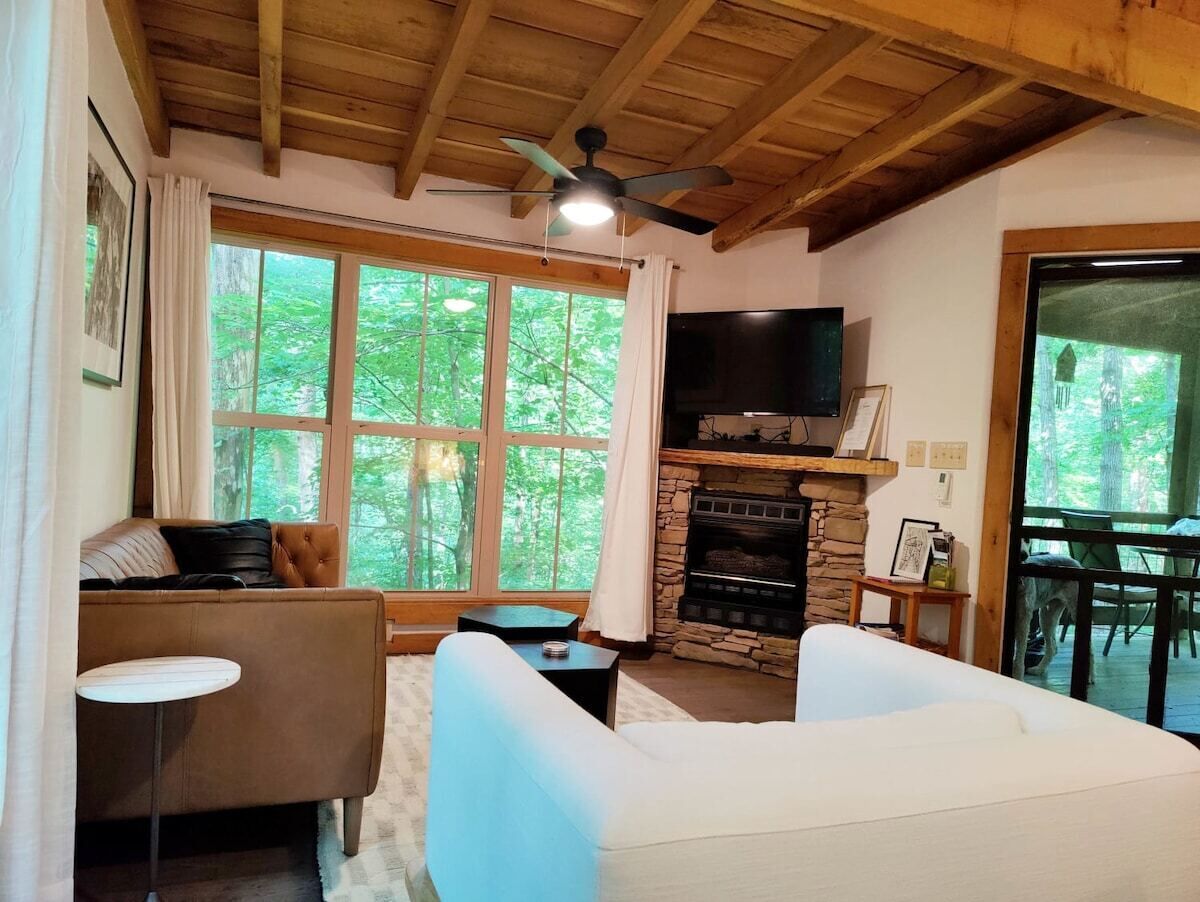 A cozy living room with a stone fireplace, a ceiling fan, and a large window overlooking a forest.