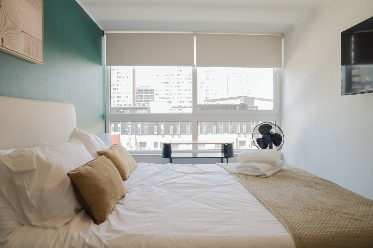 Ein Hotelzimmer mit Bett, einem Fenster mit Jalousien, einem Standventilator und Blick auf Gebäude.