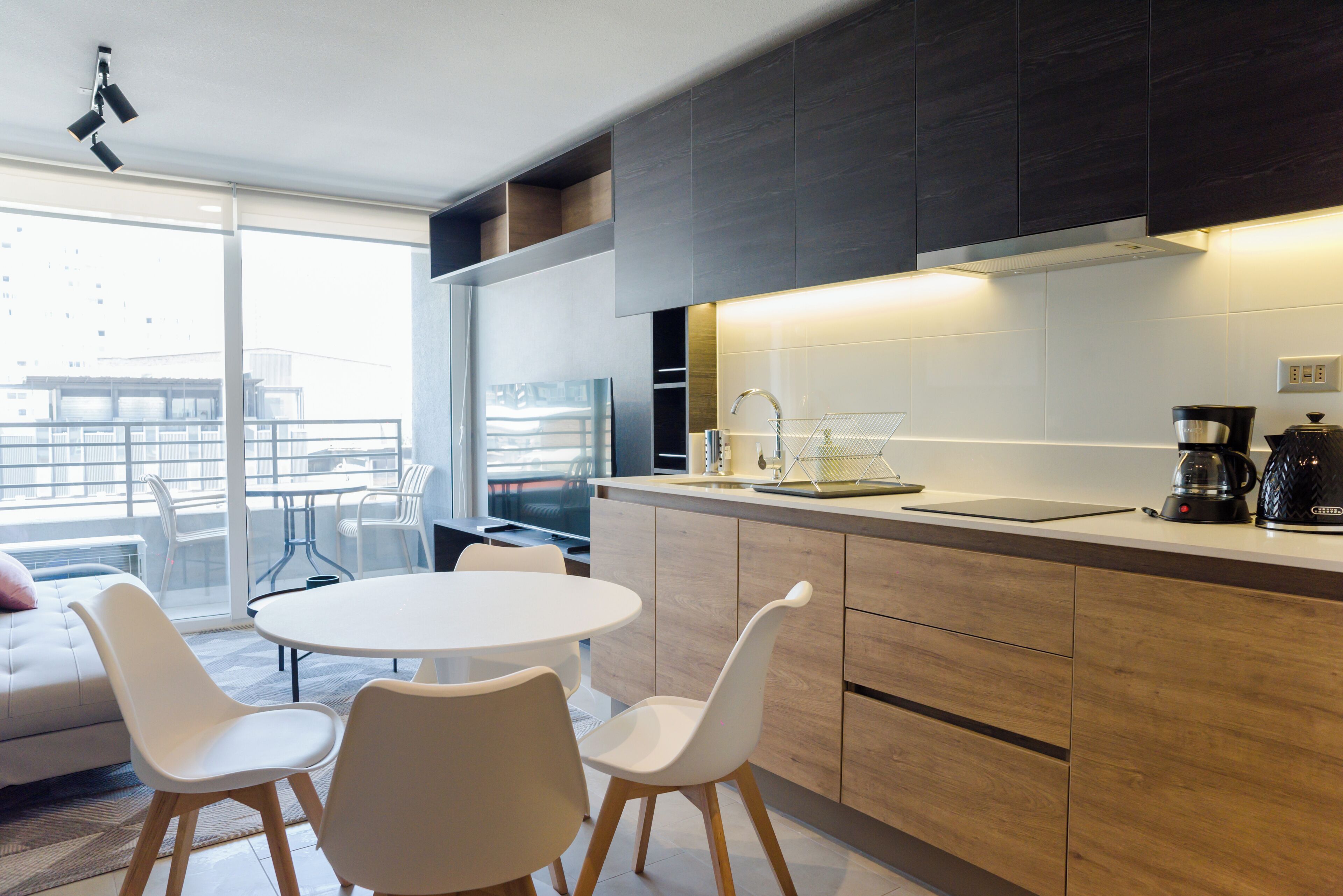 Een moderne keuken met een ronde eettafel en witte stoelen, een groot raam met uitzicht op de stad, en een balkon met een bank en stoelen.