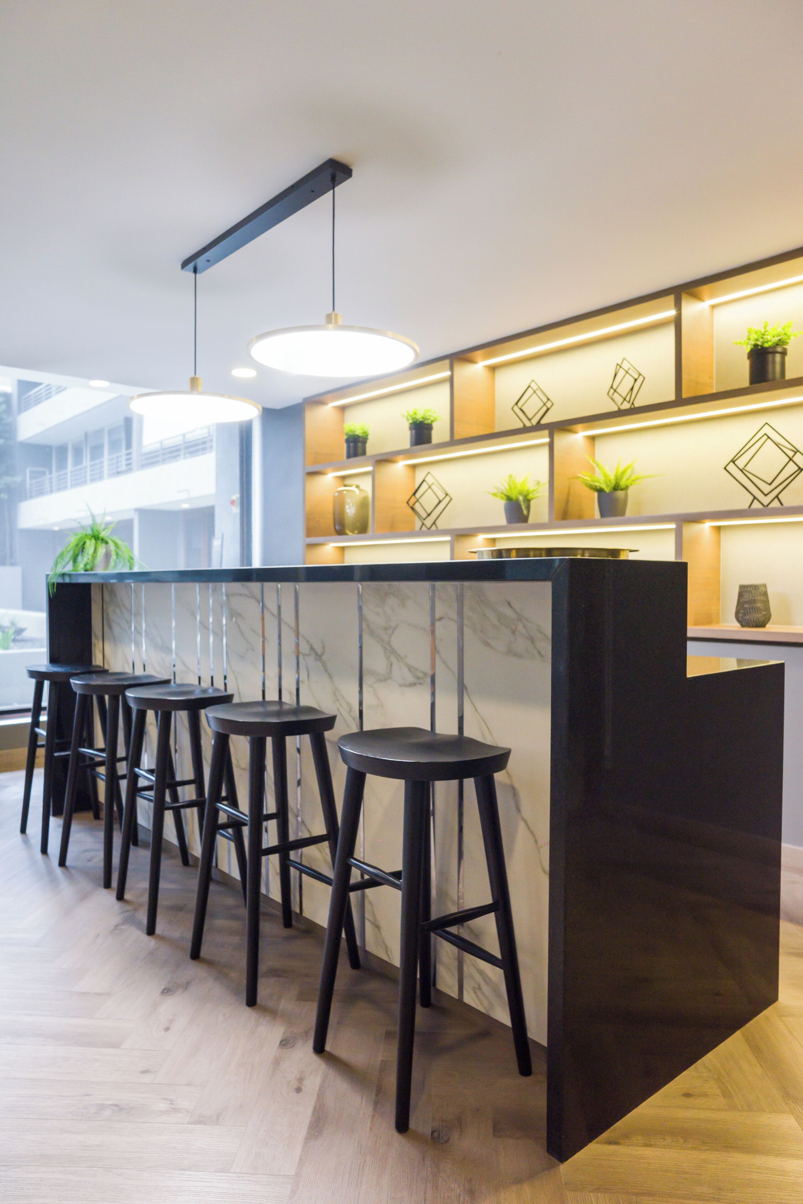 A modern bar area with a marble countertop, black stools, pendant lighting, and decorative plants.