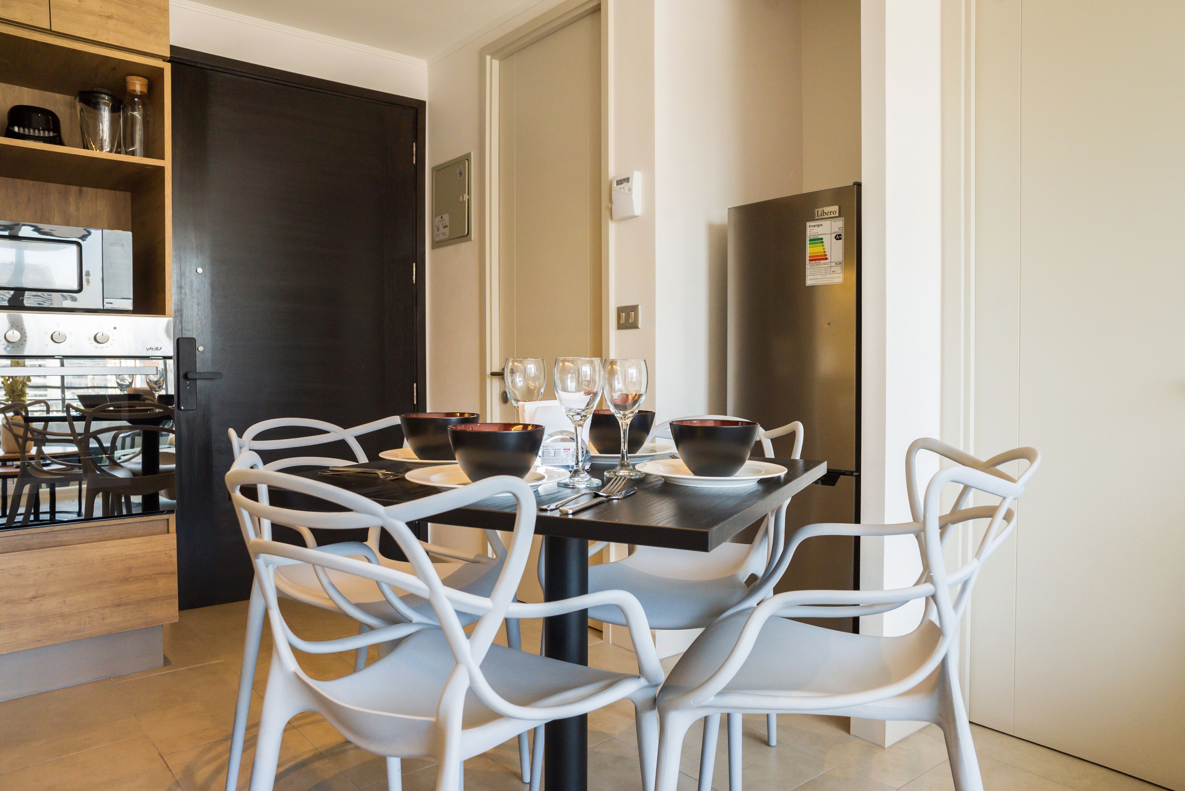 Una cocina moderna con una mesa de comedor lista para cuatro personas, sillas blancas y una encimera negra con un refrigerador.