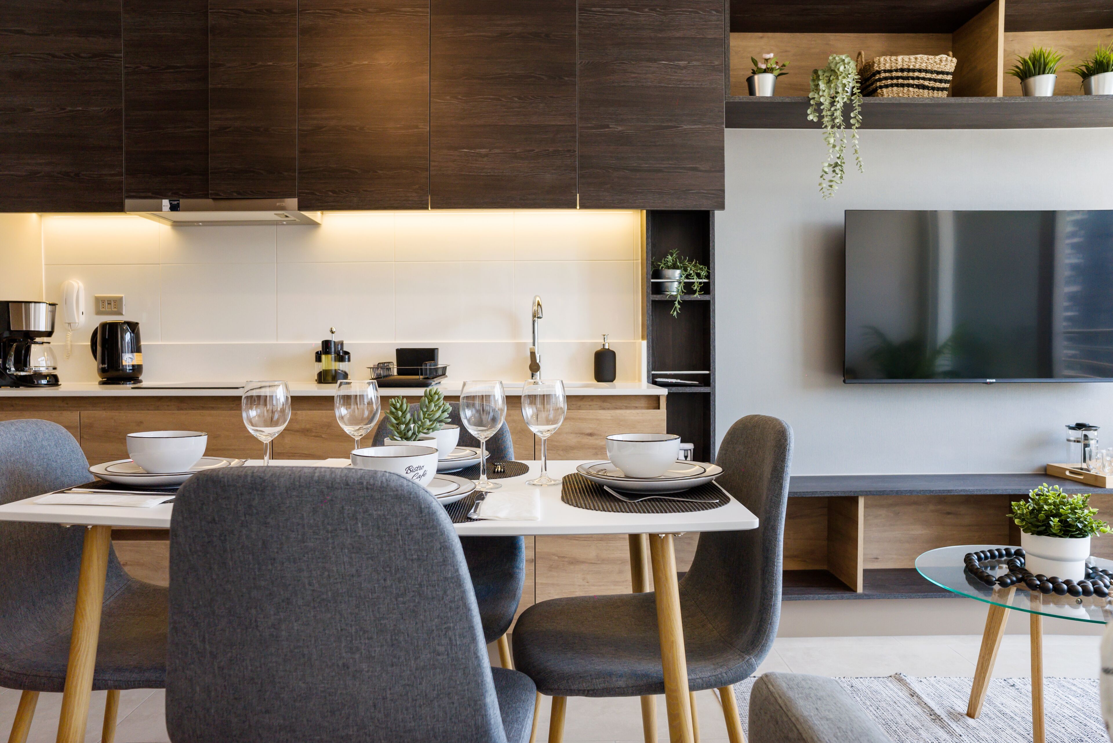 A modern kitchen with dark wood cabinets, a white countertop, and a dining area with a glass table and grey chairs.