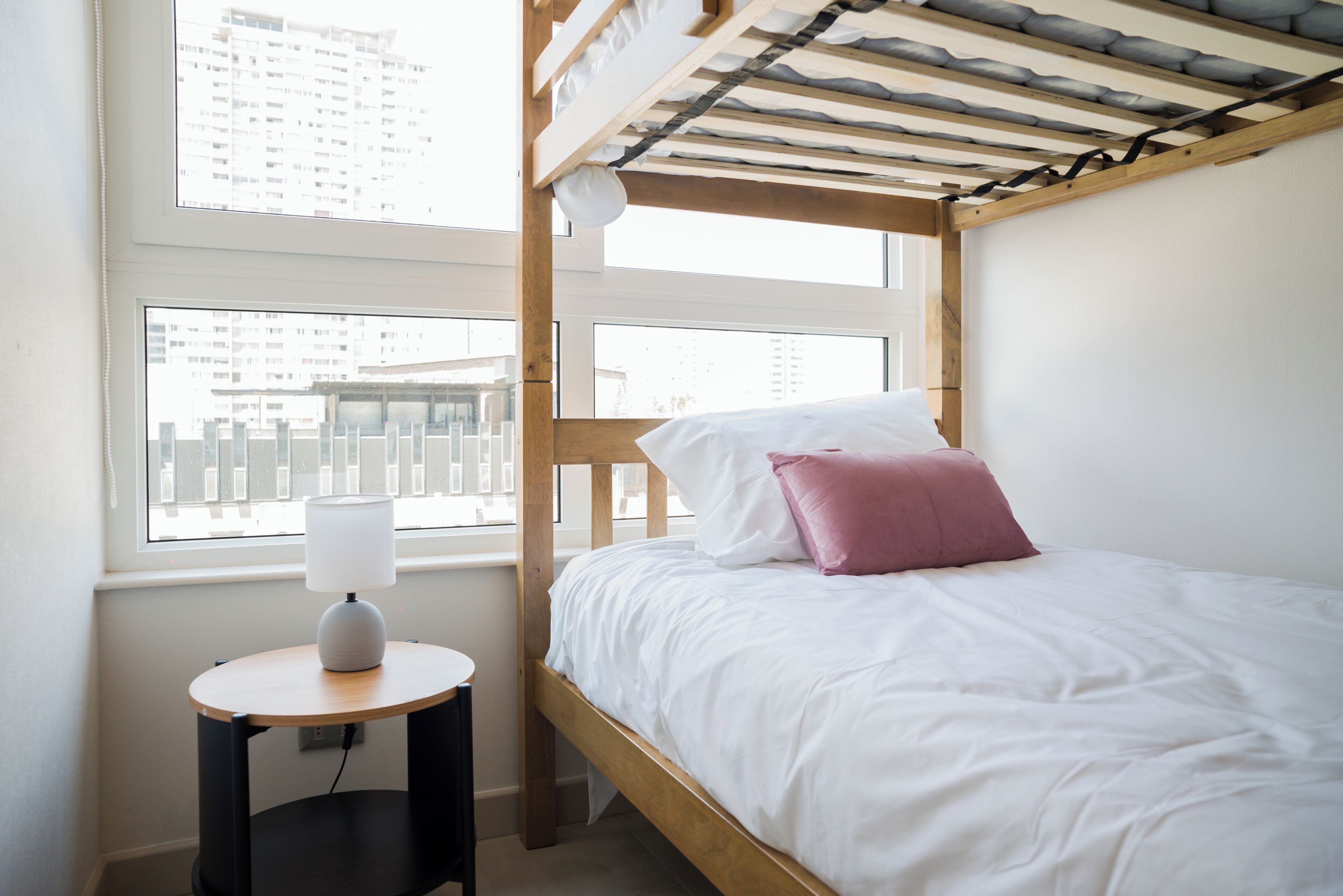 Ein Stockbett mit Holzrahmen und weißer Bettwäsche, ein Nachttisch mit Lampe sowie ein Fenster mit Blick auf Gebäude.