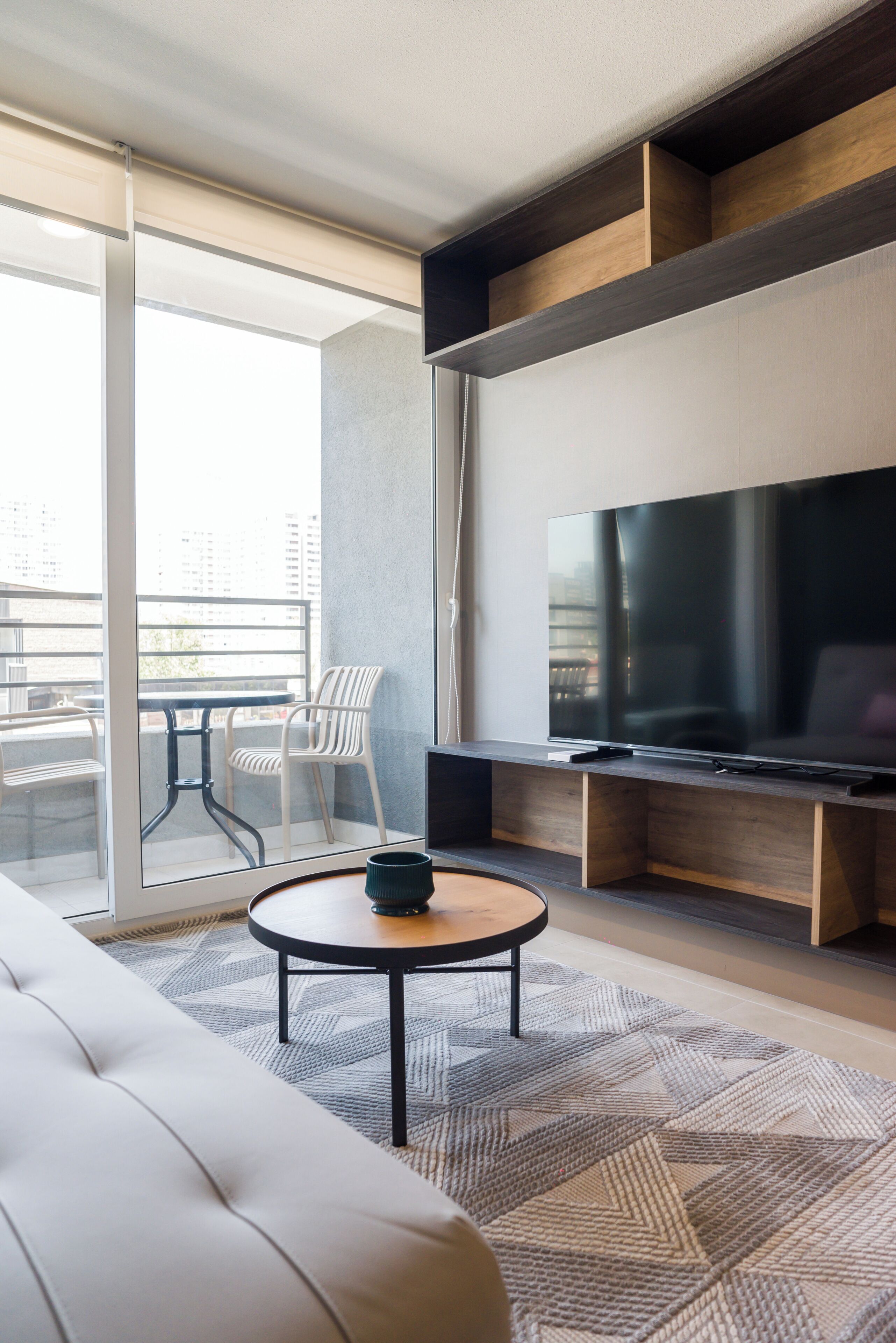 A modern living room with a flat-screen TV, a sofa, a coffee table, and a balcony with chairs.