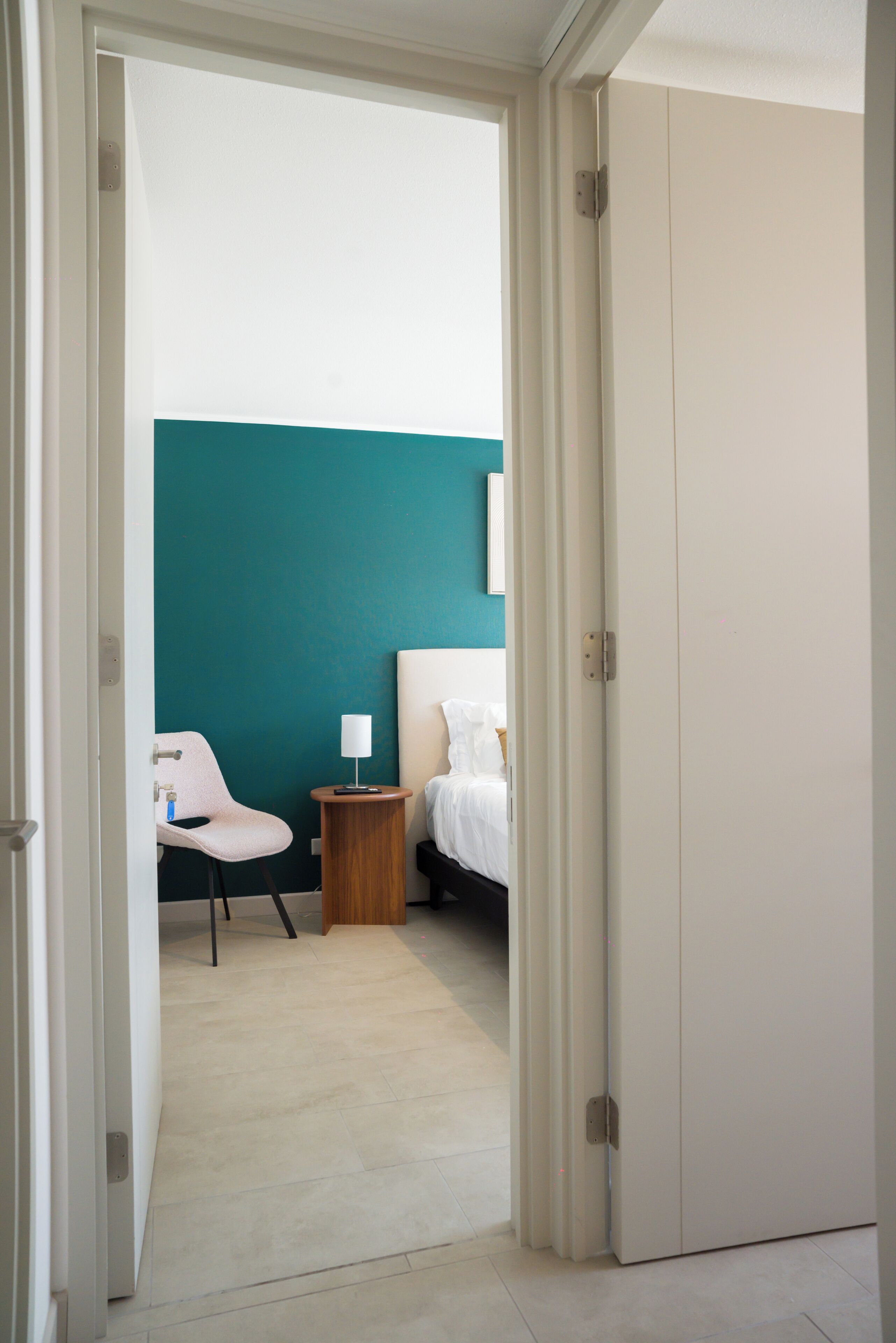 Quarto de hotel com cama, uma cadeira, mesa de cabeceira com abajur e guarda-roupa.