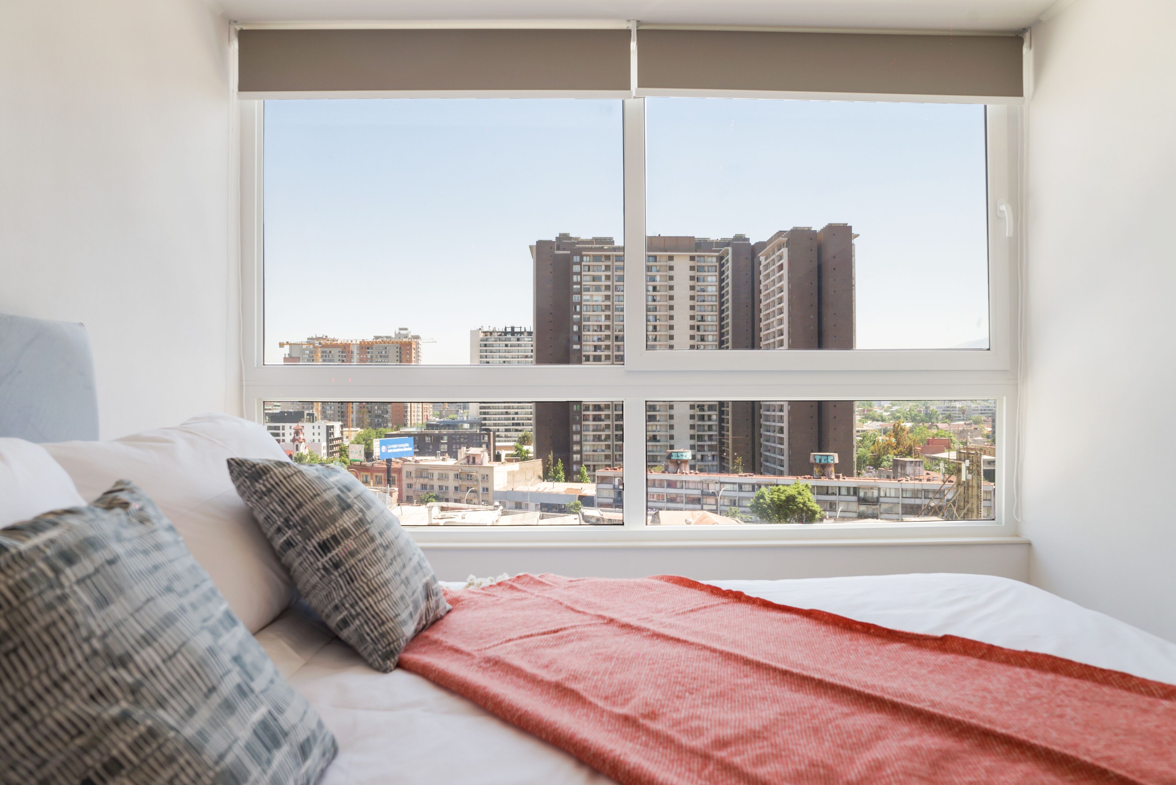 Un dormitorio con un ventanal grande, una cama con almohadas y vistas a edificios de la ciudad.