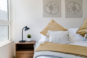 Ein Schlafzimmer mit einem Bett, Kissen, einem Nachttisch mit Lampe, einer Pflanze und einem Kunstwerk an der Wand.