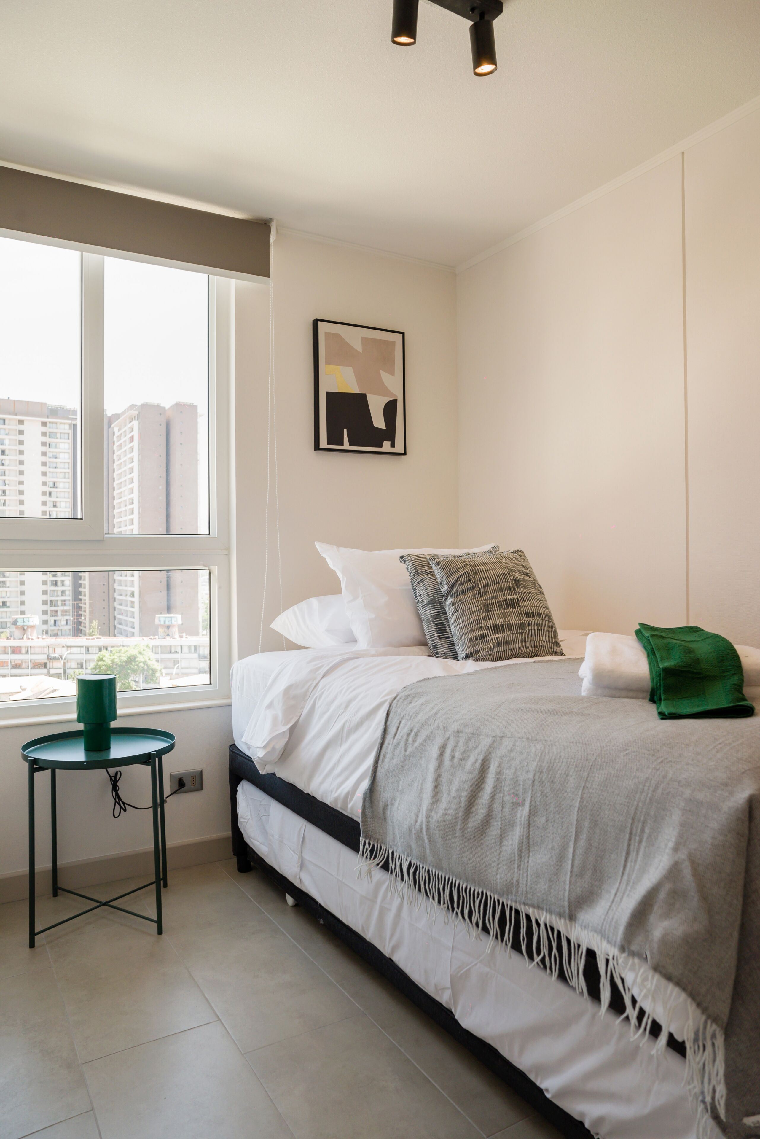 Un dormitorio con cama, una mesita, una planta verde y vistas a la ciudad a través de la ventana.