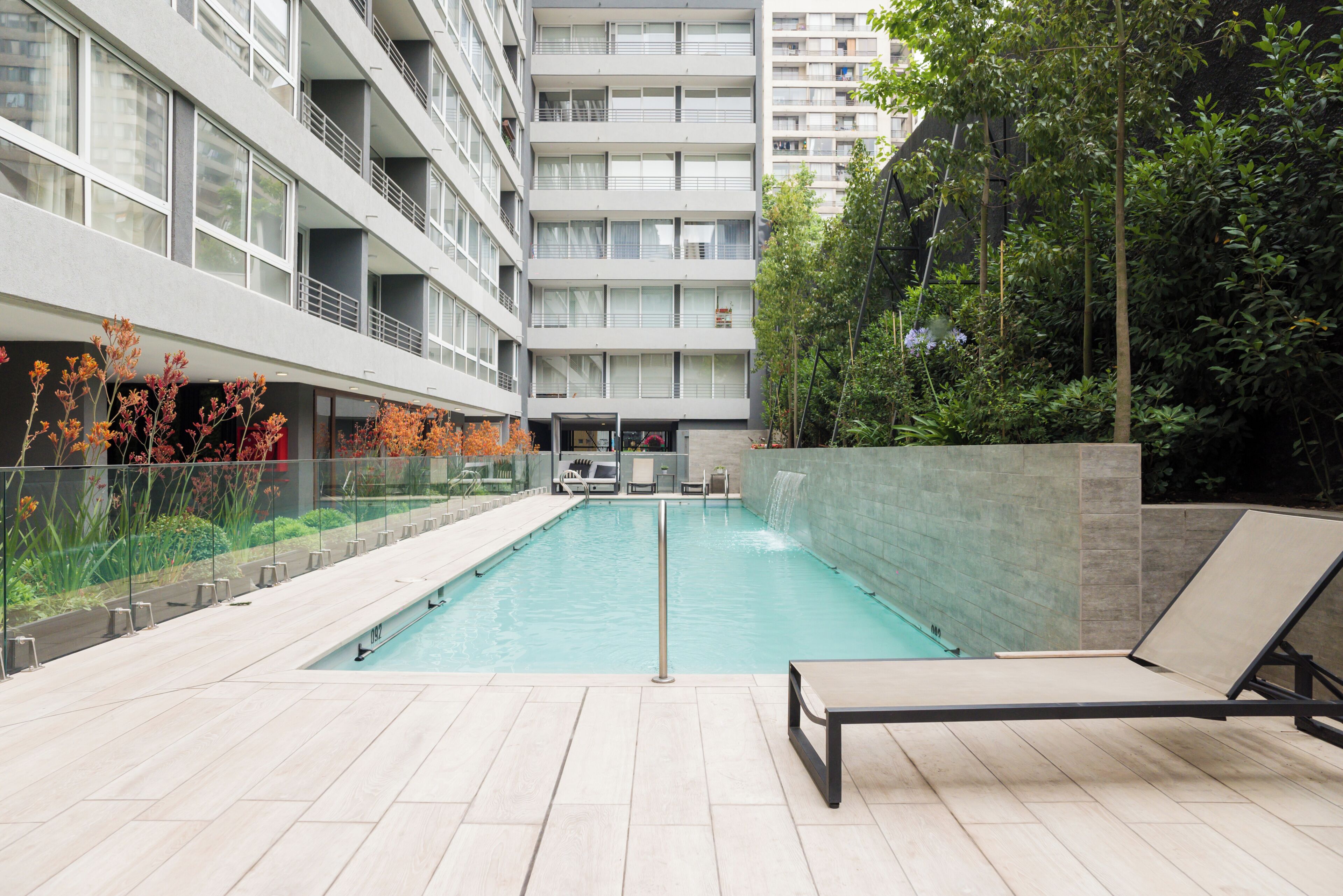 Un espace piscine extérieur moderne offrant une vue dégagée sur les immeubles environnants, avec une balustrade en verre et un fauteuil de relaxation.