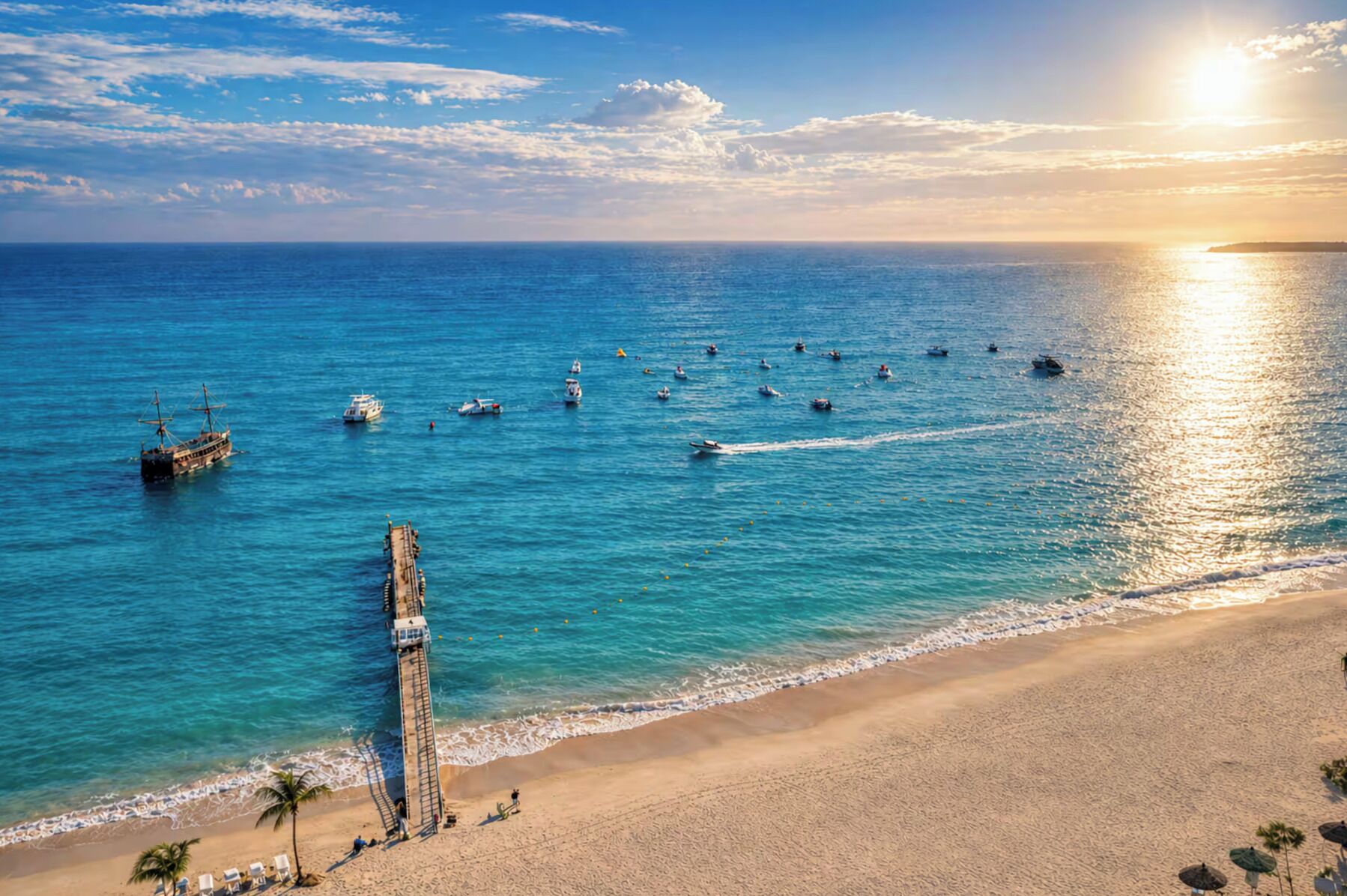 Una playa con un muelle que se adentra en el agua, rodeada de barcos y un cielo despejado.