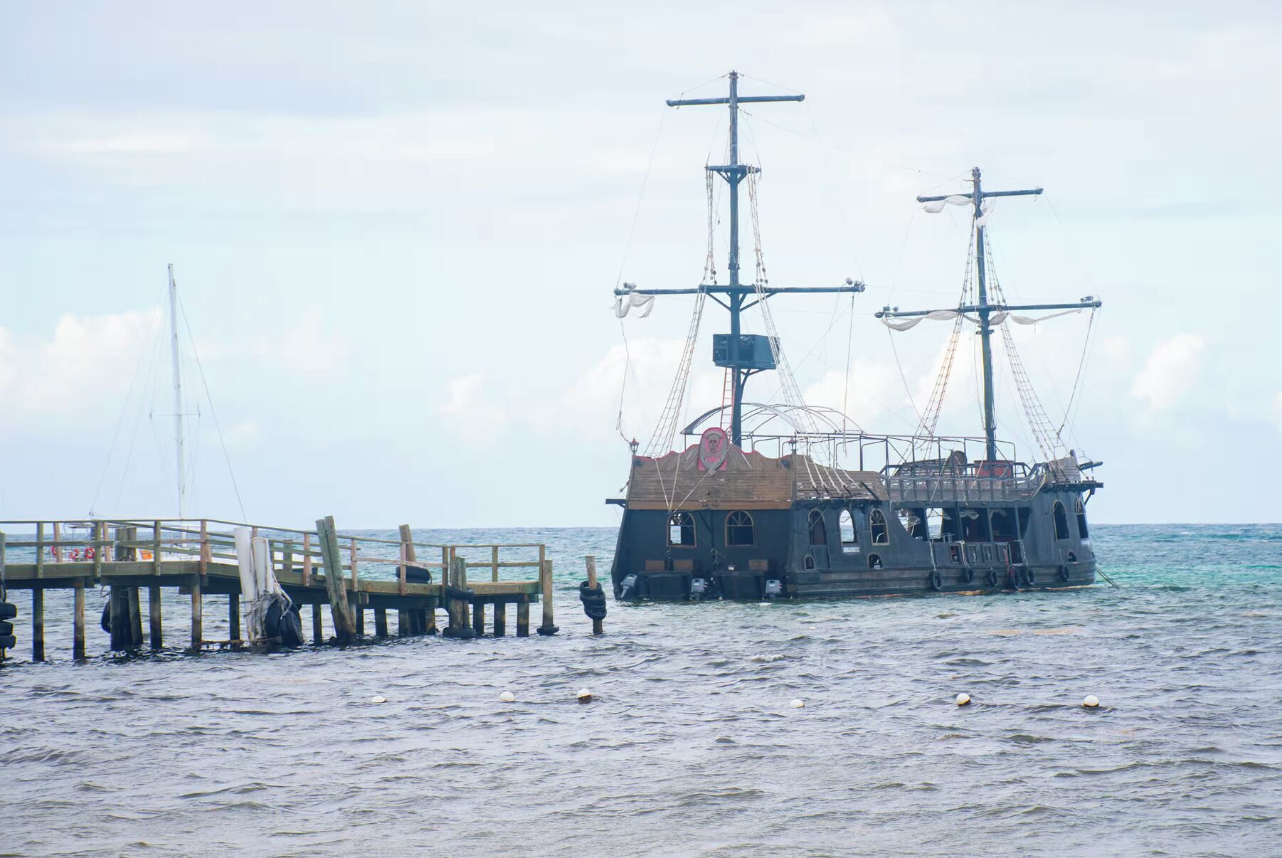 Ein Schiffswrack mit Holzboden und mehreren Masten, vor einem Steg mit einer Holzpromenade verankert.