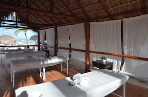 Habitación frente al mar con dos camillas de masaje, una mesita con una planta y vistas a la playa y palmeras.