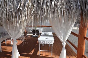 Una terraza de madera con una mesa y sillas blancas, una mesita con una planta y un techo de paja.