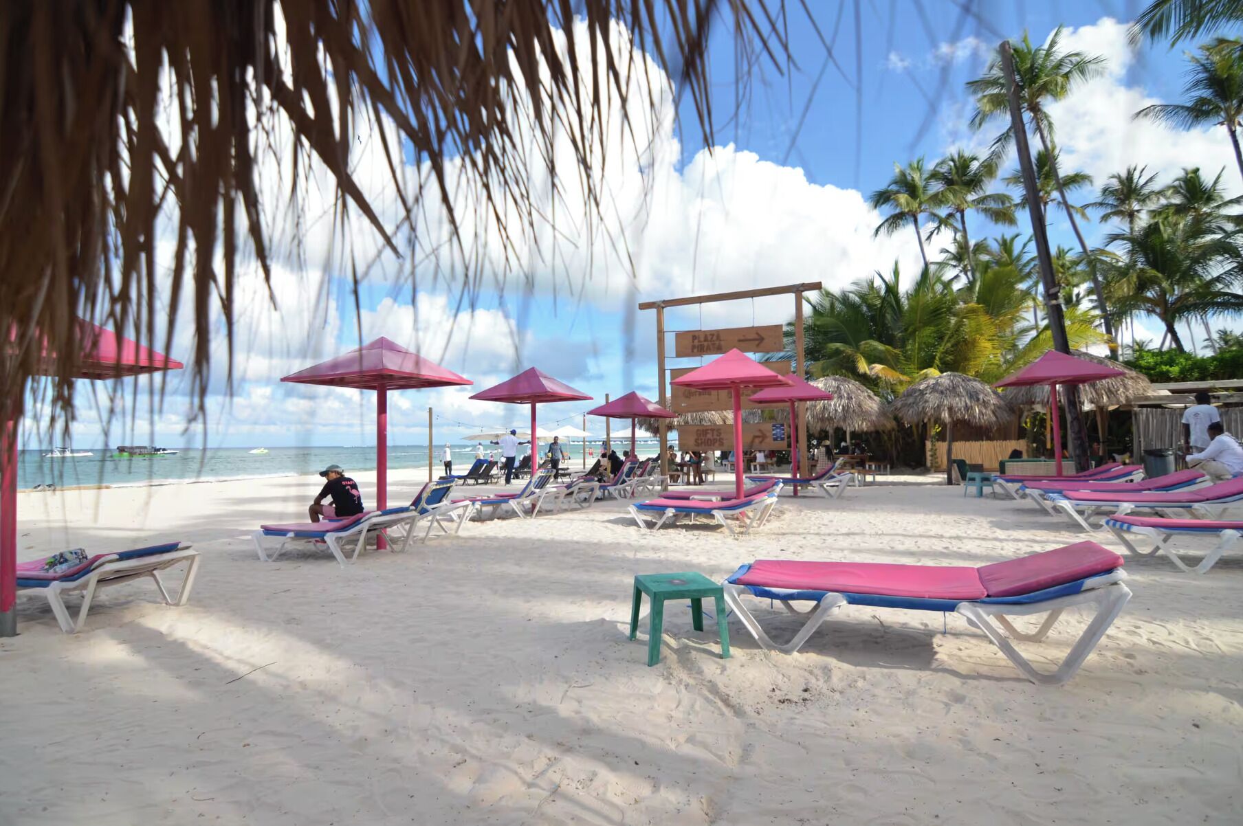 A beach with sun loungers and umbrellas, a sign for 'Plaza Pirata', and palm trees in the background.
