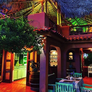 An outdoor dining area with tables and chairs, a bar area with green cabinets, and a thatched roof structure.