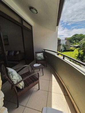 Uma varanda com mobiliário de vime, uma mesa pequena e vista para uma paisagem verde.
