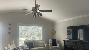 Ein Wohnzimmer mit Deckenventilator, einer Sofagarnitur, einem Fernseher und einem Fenster mit Jalousien.