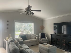 A living room with a ceiling fan, a sofa set, a television, and a window with blinds. - Welcoming  bright 3-bedroom house in sunny Naples Park. (Naples)