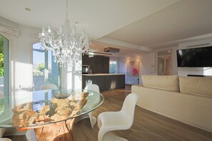 Ein modernes Wohnzimmer mit einem Glastisch, weißen Stühlen, einer Chandelüster, einer Couch und einem Fernseher.