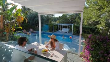 Un espace extérieur aménagé avec une véranda donnant sur la piscine, un espace de repos couvert et une végétation luxuriante.