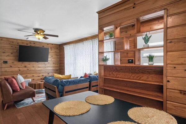 A cozy room with a sofa, a dining table with placemats, a television mounted on the wall, and a ceiling fan.
