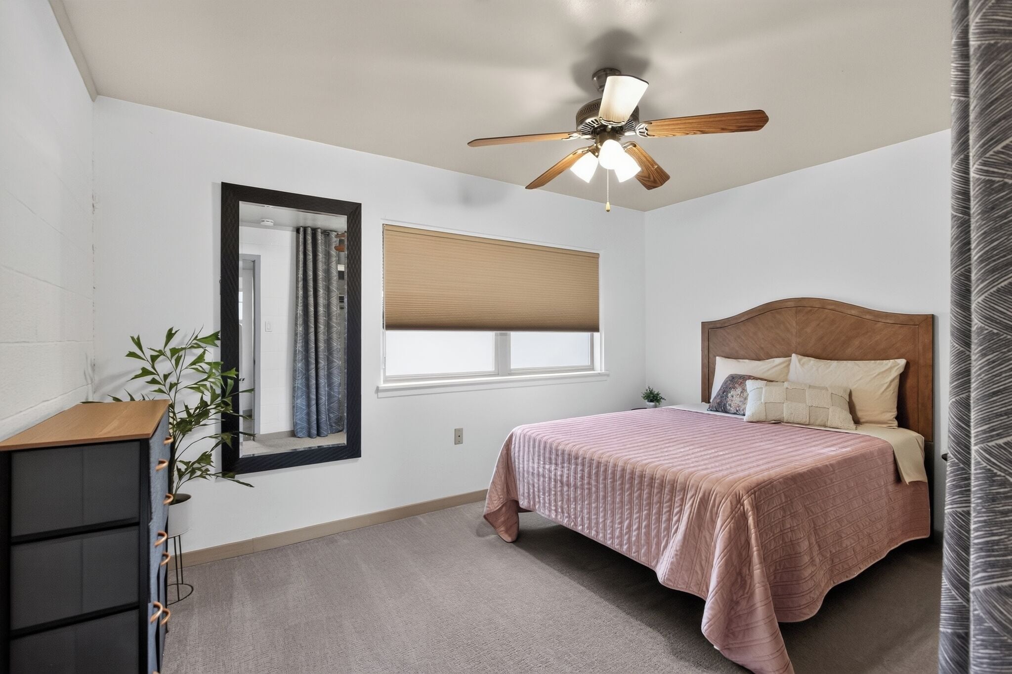 A bedroom with a bed, a ceiling fan, a window with blinds, a mirror, and a dresser.