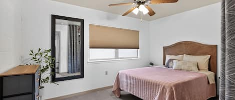 Une chambre à coucher comprenant un lit, un ventilateur de plafond, une fenêtre avec des stores, un miroir et une commode.