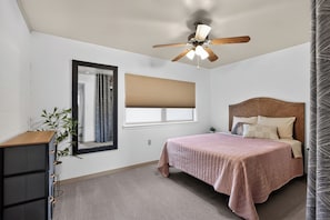 A bedroom with a bed, a ceiling fan, a window with blinds, a mirror, and a dresser.