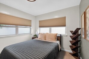 A bedroom with a bed, two windows with blinds, a bedside table with a lamp, and a wall-mounted shelf.