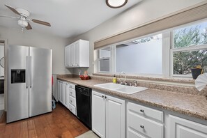 A kitchen with a built-in dishwasher, a refrigerator, a window with a view of trees, and a ceiling fan.