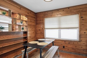 A room with wooden walls, a large window with blinds, a table with placemats, and built-in shelving with plants.