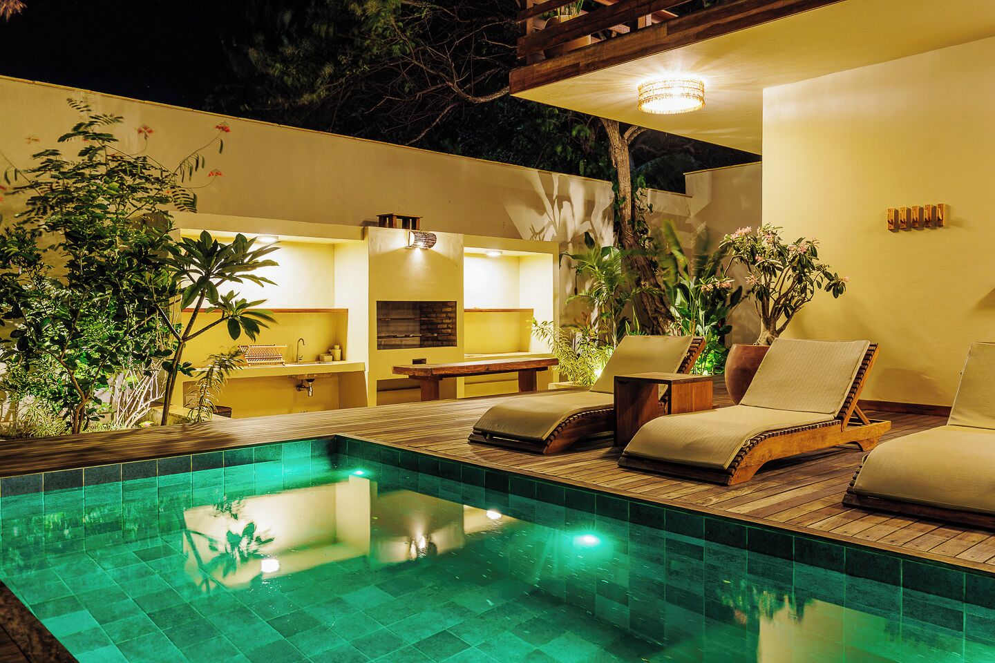 A pool area with lounge chairs and a dining table, surrounded by greenery and a yellow wall.