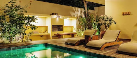 A pool area with lounge chairs and a dining table, surrounded by greenery and a yellow wall.