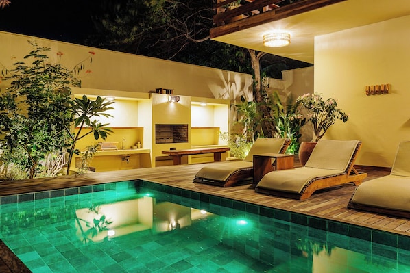 Un espace piscine avec des chaises longues et une table à manger, entouré de verdure et bordé d’un mur jaune.