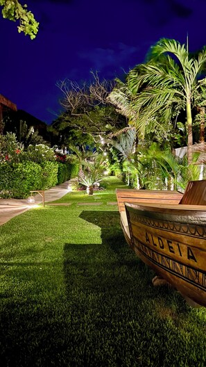 A well-lit garden with a wooden bench labeled "ALDEIA" and a pathway leading through lush greenery.