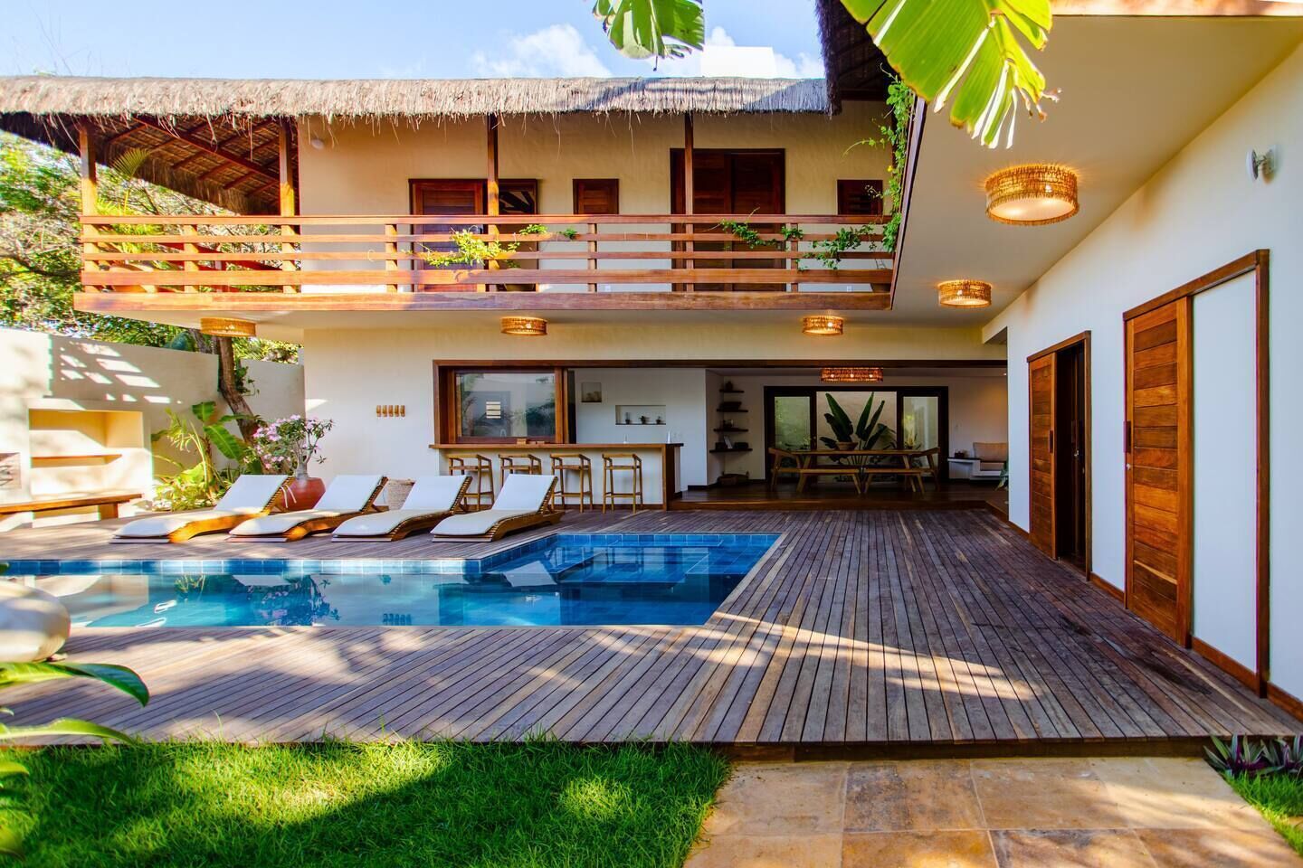 A modern villa with a pool, wooden deck, and a balcony with a thatched roof.