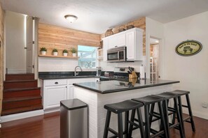 A modern kitchen with white cabinetry, a central island with bar stools, a stainless steel sink, and a microwave. - Mission Beach Bungalow w/ Direct Access to the Bay (San Diego)