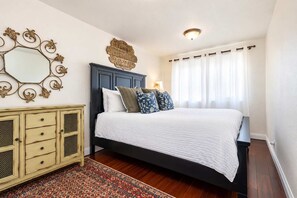 A bedroom with a dark headboard, white bedding, a patterned rug, and a decorative sign above the bed. - Mission Beach Bungalow w/ Direct Access to the Bay (San Diego)