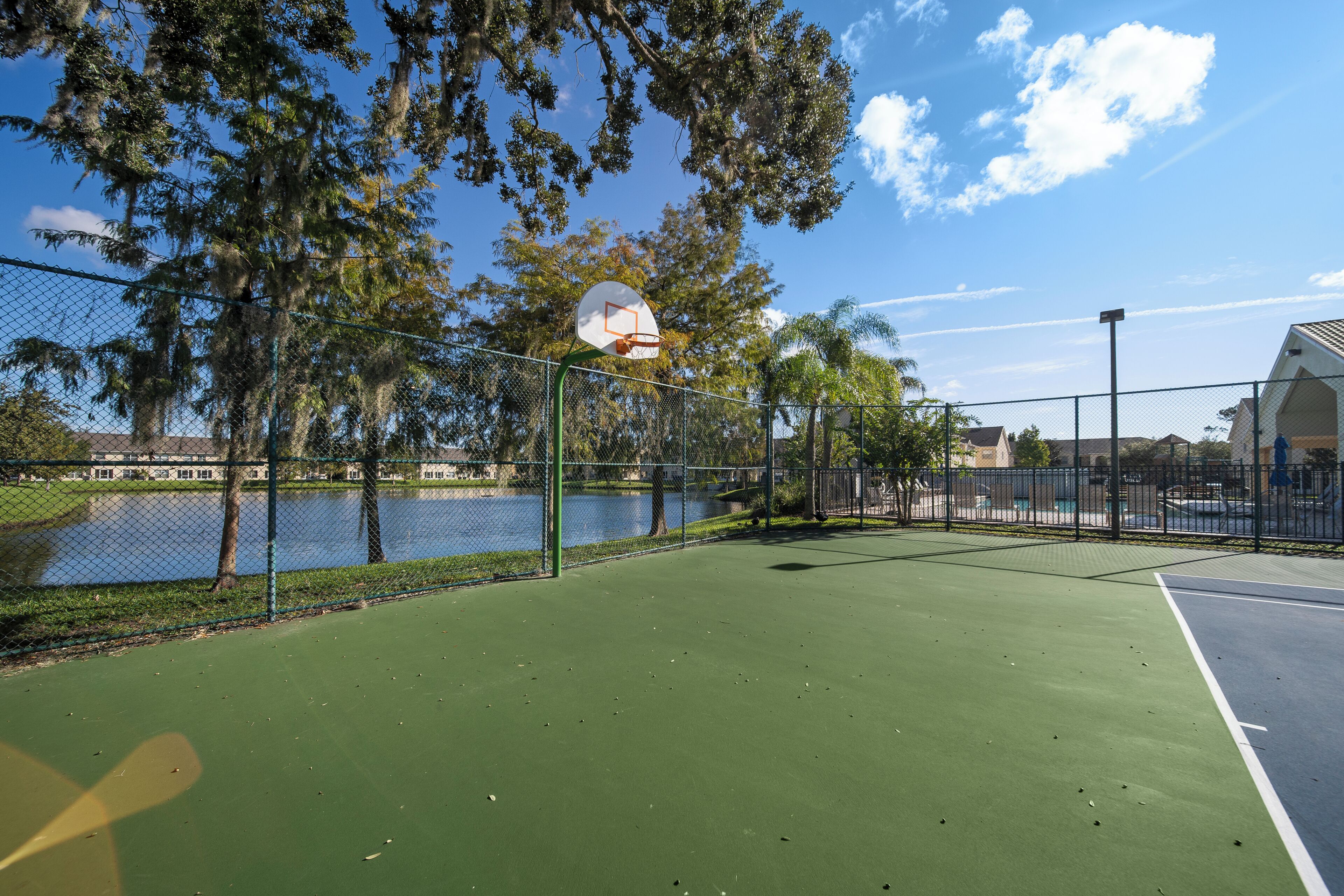 An outdoor basketball court with a chain-link fence, trees, and a body of water in the background.
