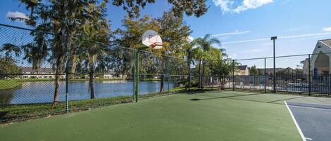 Una cancha de baloncesto al aire libre con una cerca de malla, árboles y un cuerpo de agua al fondo.