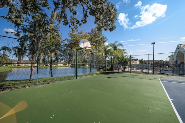 An outdoor basketball court with a chain-link fence, trees, and a body of water in the background.