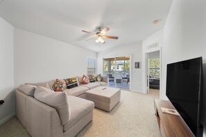 A living room with a beige sofa, ottoman, and a flat-screen TV. There is a ceiling fan, a window with blinds, and a door leading outside. - 15min from Disney - Spacious Starwars themed condo (Kissimmee)