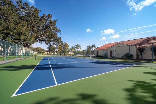 Ein Tennisplatz im Freien mit Netz, umgeben von einem Zaun, Bäumen und einem Gebäude mit rotem Dach.