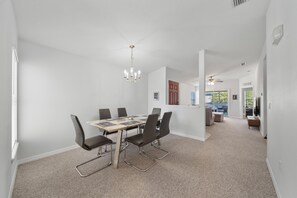 A dining area with a table, chairs, and a chandelier. There is a hallway leading to another room with a ceiling fan.