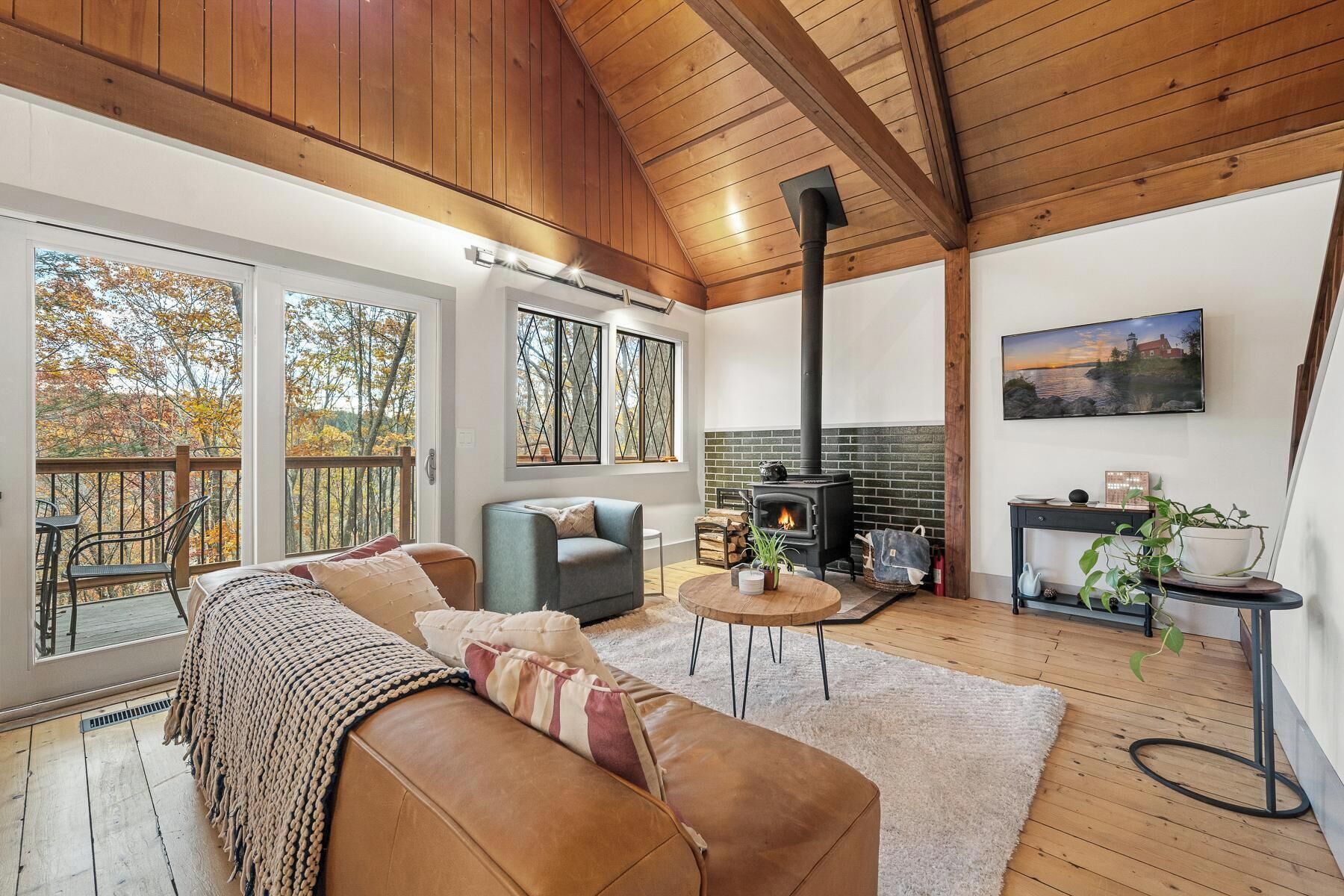 A cozy living room with a fireplace, a sofa, a coffee table, and a view of trees through large windows.