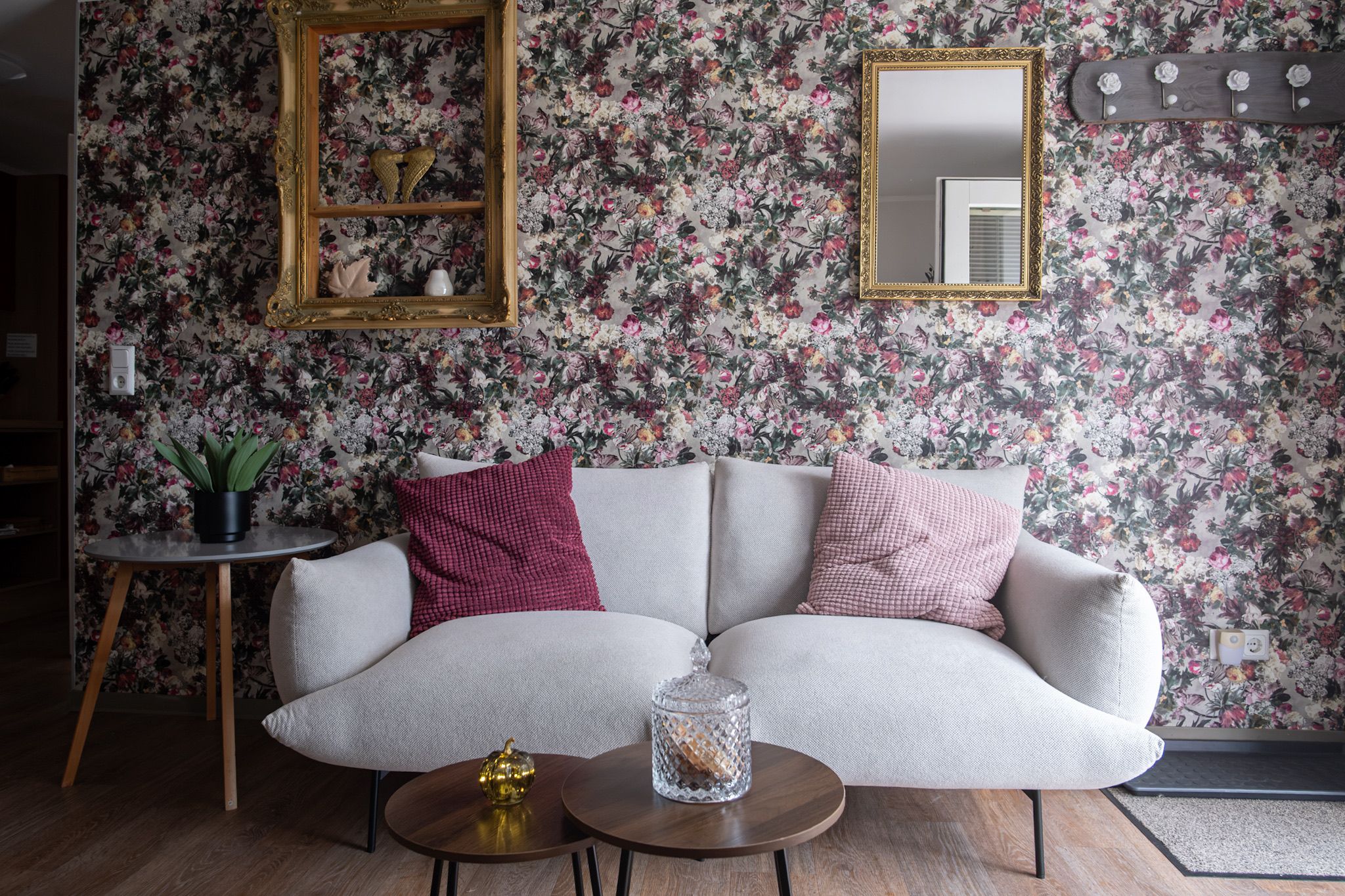 Una sala de estar con papel tapiz floral, un sofá, una mesita auxiliar y un espejo decorativo.