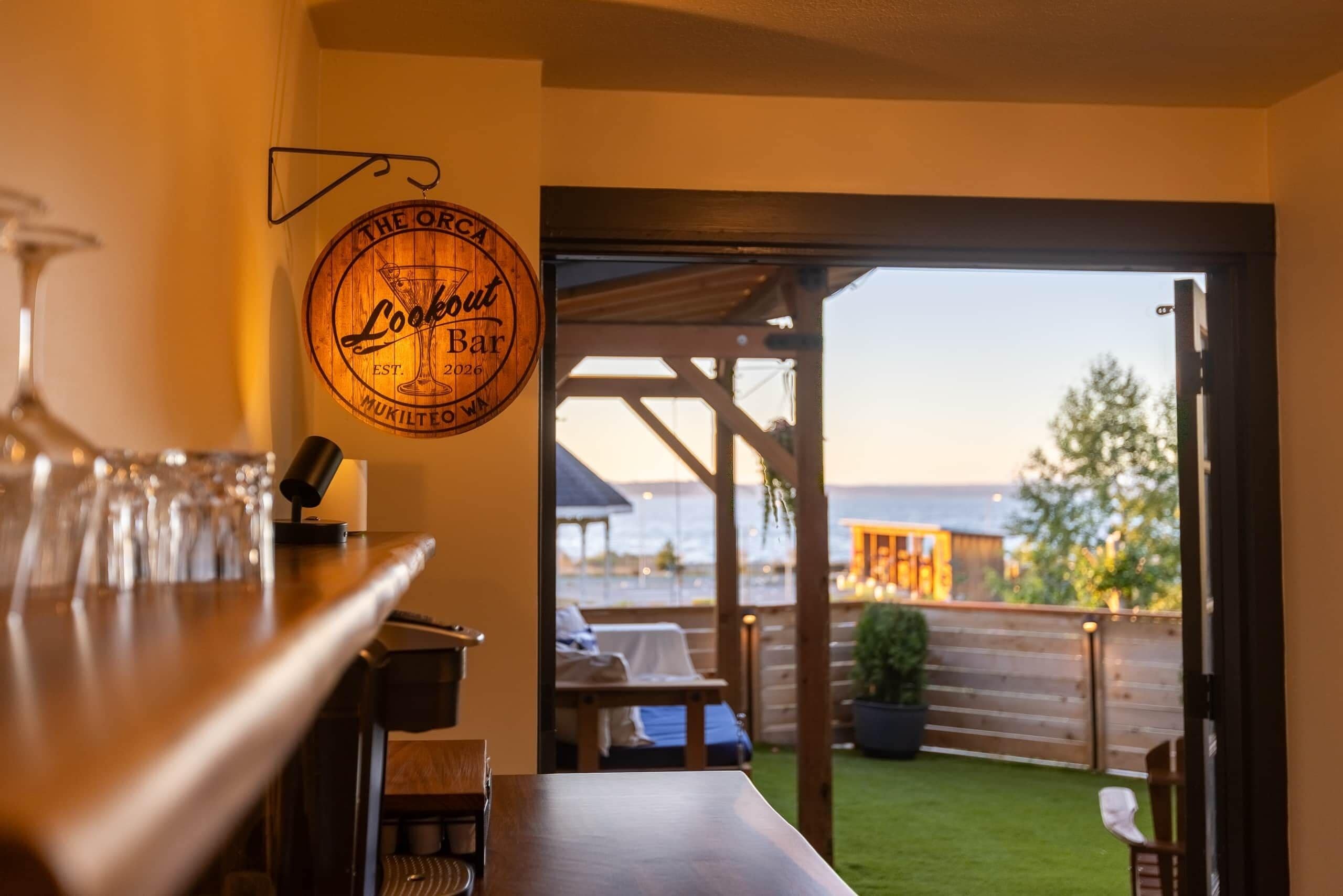 Eine Bar mit einem Holzschild, das auf ein Lokal hinweist, einem Blick auf Sitzplätze im Freien und einer Aussicht ins Grüne.