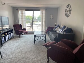 A living room with a sofa, armchairs, a coffee table, a TV, and a large window with curtains.