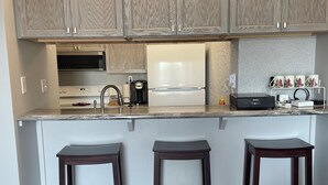 A kitchen with a built-in microwave, a refrigerator, and a coffee maker. There are three bar stools and a small shelf with cups. - Anchor Stay!  Condo w/larger deck overlooking harbor, sleeps up to 6 (Clear Lake)